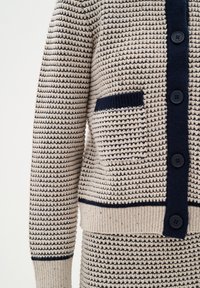 Beige knit cardigan with navy accents, textured fabric, button closure, and a single patch pocket on the left side, featuring striped patterns.
