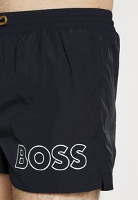 Námornícke modré plavky vyrobené z ľahkého materiálu, s elastickým pásom na šnúrku a výrazným bielym logom „BOSS“. 