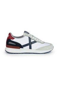 Zapatilla blanca con acentos de gamuza gris, detalles en azul marino y rojo, cierre de cordones y suela de goma texturizada. La vista lateral destaca las líneas de diseño.