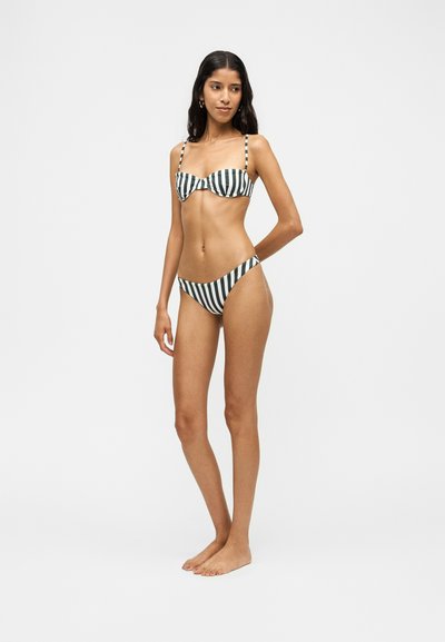 Femme debout pieds nus portant un bikini rayé vertical noir et blanc, regardant légèrement sur le côté avec un bras derrière le dos.