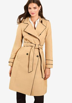 Beige trench coat featuring a double-breasted design, black piping, a belted waist, and button accents on each side. Smooth fabric texture.