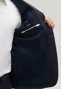 Interno di un blazer navy con tessuto liscio, una tasca etichettata, accenti di cuciture bianche e una piccola tasca funzionale.