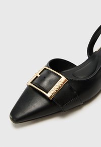 Juodas odinis slingback tipo batelis su smailiu nosiu ir auksiniu sagtimi, ant kurios užrašyta DKNY. Lygus paviršius ir minimalistinis dizainas.