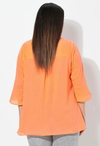Donna con capelli scuri e lisci, che indossa una blusa arancione testurizzata con maniche a 3/4 e pantaloni grigi, girata di spalle contro uno sfondo bianco.