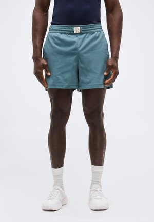 Άνδρας που φοράει πετρόλ αθλητικό σορτς, λευκές καλτσοδέτες και λευκά αθλητικά παπούτσια, στέκεται μπροστά σε απλό ανοιχτό φόντο.