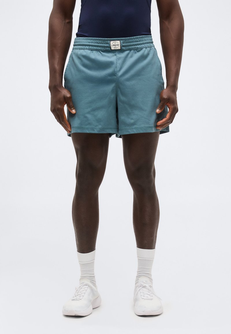 Άνδρας που φοράει πετρόλ αθλητικό σορτς, λευκές καλτσοδέτες και λευκά αθλητικά παπούτσια, στέκεται μπροστά σε απλό ανοιχτό φόντο.