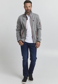 Grå fleecjakke med oransje detaljer, glidelåslommer, høy krage og en teksturert overflate. Båret over en hvit t-skjorte og blå jeans.