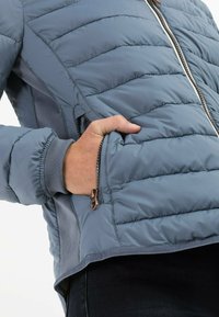 Lyseblå pufferjakke med vandret syninger, lynlåslommer og sidelæg i en mørkere farve. Ribbede manchetter forbedrer pasformen.