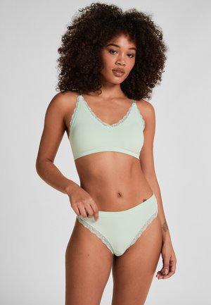 Donna con capelli ricci che indossa un completino di lingerie verde chiaro a coste con bordi in pizzo, in piedi davanti a uno sfondo semplice grigio chiaro.