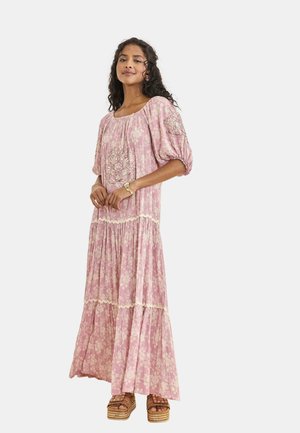 Mujer con cabello oscuro y rizado, vistiendo un vestido largo de varias capas con flores rosas y sandalias con plataforma, de pie con las manos delicadamente entrelazadas.