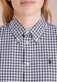 Svartvitt gingham-mönstrad skjorta med knappar och krage, med vita knappar och en liten broderad logotyp på vänster bröst.