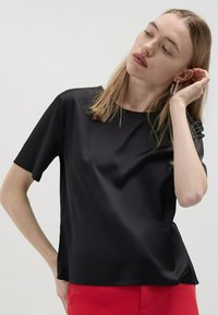 Blusa nera a maniche corte in tessuto liscio con vestibilità rilassata, caratterizzata da un collo rotondo e una leggera lucentezza. Abbinata a pantaloni rossi.