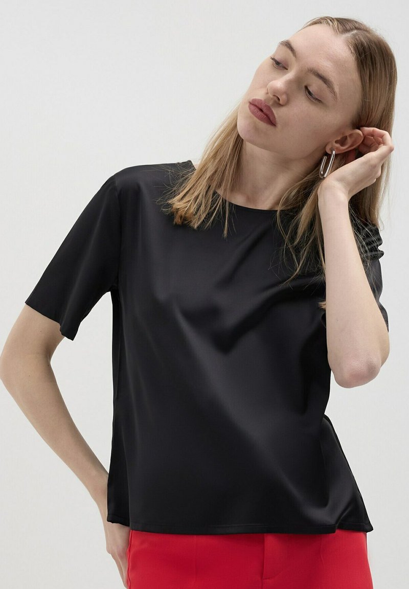 Blusa nera a maniche corte in tessuto liscio con vestibilità rilassata, caratterizzata da un collo rotondo e una leggera lucentezza. Abbinata a pantaloni rossi.