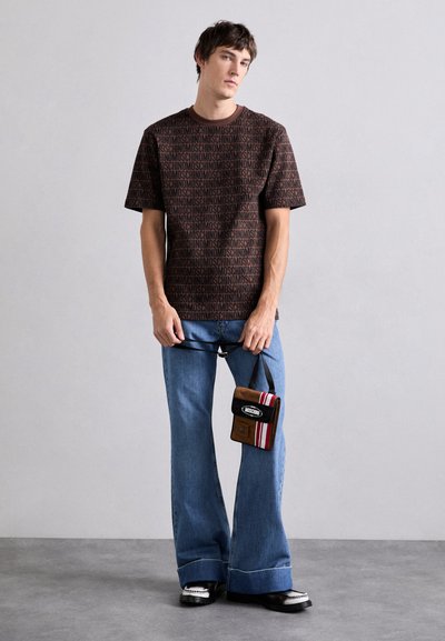 Tricou graphic maro cu model "MOSCHINO", asortat cu blugi evazați albaștri, purtând o geantă marron cu accente negre și roșii.