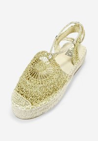 Espadrilles de encaje dorado con un diseño tejido, forro de lona color crema y suela de yute. Presenta cortes decorativos y correas ajustables.
