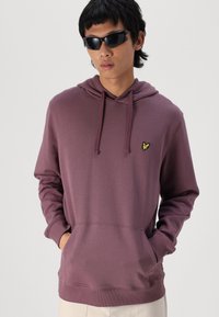Sudadera morada hecha de tela suave, con un bolsillo canguro y un pequeño logo amarillo en el pecho. El diseño incluye una capucha clásica.