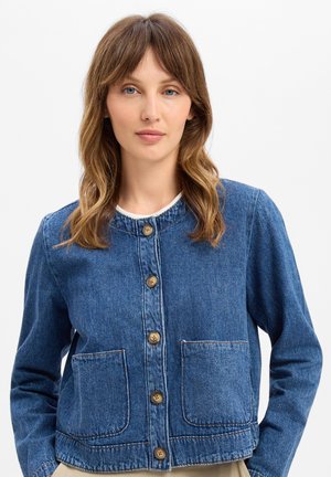 Vrouw met bruin haar tot op de schouders draagt een blauw denim jasje met knopen en twee zakken aan de voorkant tegen een effen witte achtergrond.