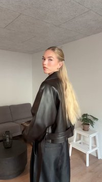 Svart läderjacka med skärp i midjan och bred krage, med en slät yta. Långt blont hår stylat bakåt med en slät frisyr.