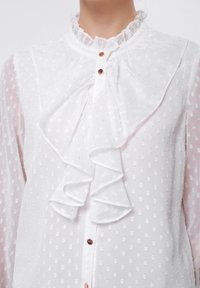 White blouse with a ruffled collar, textured fabric featuring small dotted patterns, and decorative button closures in the front.