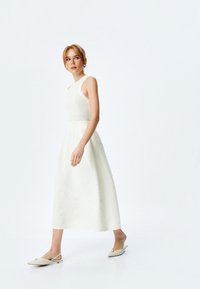 Robe blanche avec un corsage ajusté et une jupe texturée et brodée. Portée avec des chaussures à talons ouvertes beige présentant un accent décoratif.