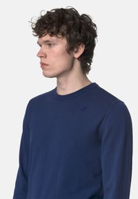 Maglione lavorato a maglia blu navy con scollatura rotonda e maniche lunghe. Presenta un logo ricamato discreto sul petto. Texture liscia, design aderente.
