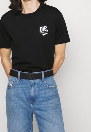 Personne portant un T-shirt Diesel noir rentré dans un jean bleu taille haute avec une ceinture en tissu noire, les mains derrière le dos.