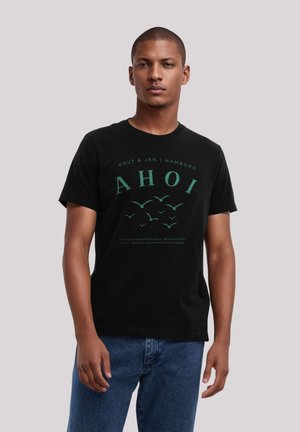 Black cotton t-shirt featuring a green graphic with the word "AHOI" and bird illustrations, combined with text about coastal life.