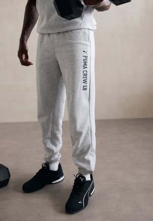 Grijze joggingbroek met golfboorden aan de enkels, voorzien van "PUMA CREW 48" in marineblauw op de zijkant; gecombineerd met zwarte sportieve schoenen.