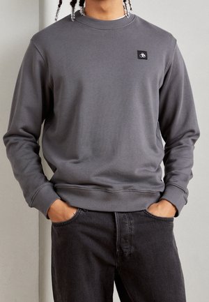 Osoba nosząca gładką szarą bluzę z okrągłym dekoltem z małym czarno-białym logo na piersi, z rękami włożonymi do kieszeni czarnych dżinsów.