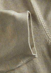 Beżowy sweter dzianinowy z detalami, pokazujący ribbowe mankiety i gładką strukturę, z subtelnymi odmianami koloru. Posiada czyste akcenty szwów.