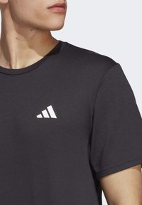 Close-up of person wearing black Adidas t-shirt showing white logo on chest and part of neck, jaw, and arm against plain background.