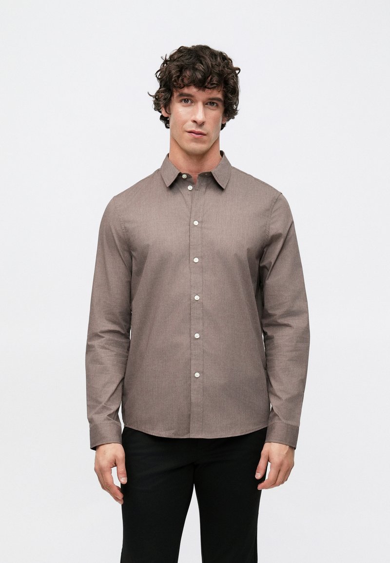 Braunes Hemd mit Knopfleiste aus strukturiertem Stoff, mit spitzem Kragen, langen Ärmeln und weißen Knöpfen. Kombiniert mit schwarzer Hose.