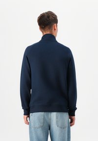 Hanorac bleumarin cu guler înalt, cu manșete și tiv elastic, din material moale, prezentat din spate, asortat cu blugi albaștri deschis.