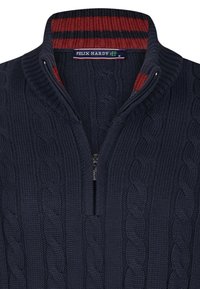 Granatowy sweter dzianinowy z wzorem w warkocze, z zapięciem na pół-zamek z przodu oraz czerwoną paskowaną podszewką kołnierza. Posiada metkę z logo marki na szyi.