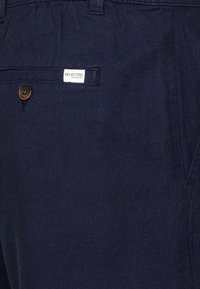 Gros plan d'une poche de pantalon bleu marine avec un bouton marron et une petite étiquette blanche portant la mention "SELECTED HOMME."