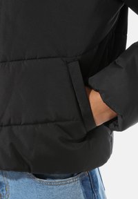 Czarna pikowana kurtka puchowa o gładkiej fakturze i przeszytym wzorze, z zewnętrzną kieszenią na wysokości talii, zestawiona z jasnoniebieskimi dżinsami.