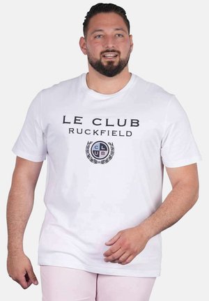 Homme aux cheveux foncés et à la barbe portant un t-shirt blanc « Le Club Ruckfield » et un pantalon clair, debout devant un fond uni.