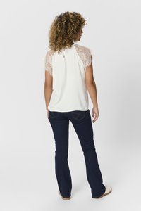 Top bianco con maniche in pizzo e dettaglio a oblò sulla schiena, abbinato a jeans a zampa d'elefante blu scuro e scarpe chiare. Tessuto morbido, design casual.