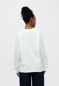 Άσπρο βαμβακερό πουκάμισο με μακριά μανίκια, χαλαρή εφαρμογή, πέτσα ώμων και στρογγυλή λαιμόκοψη. Ορατή ραφή κατά μήκος των μανσετών και του τελειώματος.