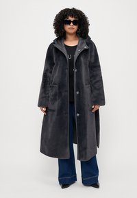 Manteau en fausse fourrure gris foncé, long, avec une capuche, des boutons à l'avant et des poches latérales. Texture lisse, coupe ample et manches larges. Convient pour le superposition.
