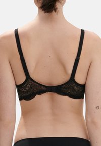 Reggiseno in pizzo nero con spalline regolabili, bordi ondulati e una chiusura decorativa sul retro. Il pizzo presenta motivi e texture intricate.