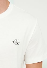 Biele bavlnené tričko s okrúhlym výstrihom. Na ľavej strane na hrudi je čierne logo „CK“ a nápis „Calvin Klein Jeans“. Krátke rukávy.