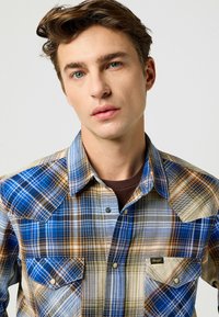 Camisa de cuadros abotonada en tonos azules, marrones y beige; con dos bolsillos en el pecho; yugo de estilo western; tejido de algodón con una textura suave.