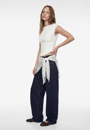 Frau trägt ein ärmelloses weißes Oberteil, weite dunkelblaue Jeans, einen weißen Spitzen-Schal, der an der Taille gebunden ist, und dunkelbraune Ballerinas mit Nieten.