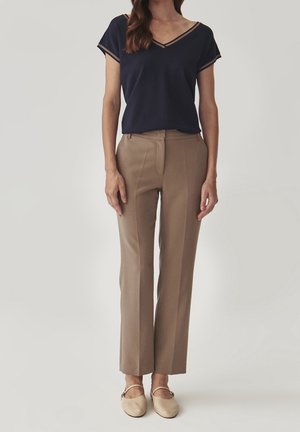 Frau steht und trägt ein marineblaues, kurzärmliges V-Ausschnitt-Oberteil, beiges gerade geschnittenes Hosenbein und beige Mary-Jane-Ballerinas.