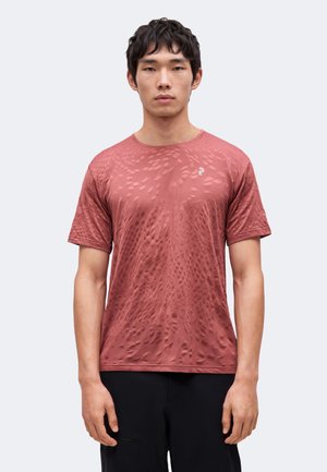 Jeune homme portant un t-shirt à manches courtes texturé bordeaux et un pantalon noir, debout devant un fond clair uni.