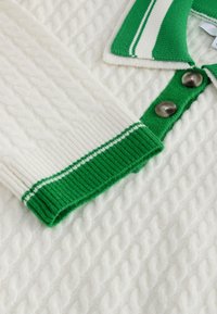 Pull tricot cable blanc avec col et poignets à rayures vertes et blanches, avec deux boutons marron près du col.