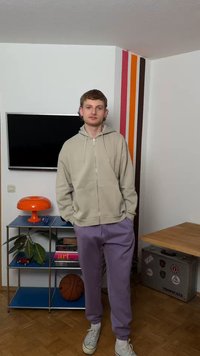 Νέος άνδρας με μπεζ φούτερ με κουκούλα και μωβ φόρμα στέκεται σε δωμάτιο με μπασκετόσφαιρα, πορτοκαλί λάμπα, τηλεόραση και ριγέ διακοσμητικό στον τοίχο.