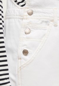 Λευκό τζιν με τρία μεταλλικά κουμπιά στην άκρη της τσέπης, δίπλα σε ύφασμα με ασπρόμαυρες ρίγες.