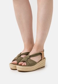 Olive green espadrille sandals with knotted fabric straps, a woven jute platform, and a natural rubber sole. Adjustable ankle strap detail.